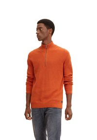 Pull-over orange en tricot zippé avec une texture côtelée, manches longues et un col montant. Associé à un jean bleu, avec un petit détail de logo.