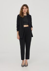 Giacca da abito nera su misura con un top nero corto, pantaloni neri slim-fit e tacchi in argento texturizzati, su uno sfondo bianco.