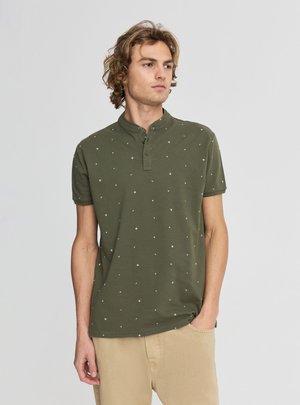 Giovane uomo con capelli ricci che indossa una camicia verde oliva a maniche corte con piccoli motivi bianchi e pantaloni beige con una mano in tasca.