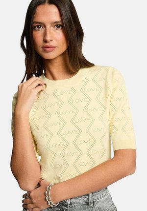 Femme aux longs cheveux foncés portant un haut en maille jaune pâle avec le motif "LOVE" et un jean délavé clair, posant avec la main près du visage.