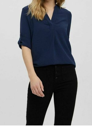 Vrouw draagt een marineblauwe blouse met lange mouwen die zijn opgerold, en een zwarte broek met hoge taille, knopen aan de voorkant en zakken.