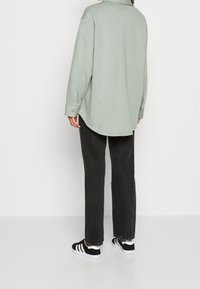 Camicia oversize button-up verde chiaro con orlo arrotondato, abbinata a jeans grigio scuro e scarpe nere con strisce bianche.