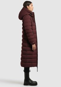 Burgunderfarbener Puffermantel mit einer langen Silhouette, Kapuze und schwarzen Reißverschlüssen. Ein gestepptes Design sorgt für eine strukturierte Optik. Kombiniert mit schwarzen Stiefeln.