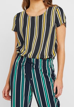Femme portant un t-shirt à manches courtes rayé avec des lignes jaunes et blanches, et un pantalon à cordon rayé avec des lignes vertes, blanches et bleu marine.