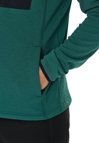 Camicia verde a maniche lunghe realizzata in tessuto testurizzato, con una tasca nera e polsini a contrasto, evidenziando una vestibilità comoda e un design pratico.