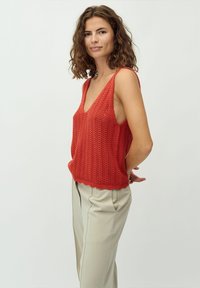 Rotes gestricktes Tanktop mit V-Ausschnitt und offenen Seiten, kombiniert mit hellbeigen Hosen mit Falten. Einfaches, strukturiertes Design.