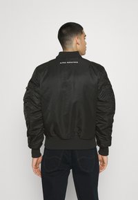 Giacca bomber nera con una texture liscia e lucida, colletto e polsini a costine, con il logo "ALPHA INDUSTRIES" sul retro.