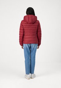 Rød pufferjakke med horisontale sømmer, festet hette, elastiske mansjetter og en tilpasset midje. Kombinert med blå denimjeans og hvite sneakers.