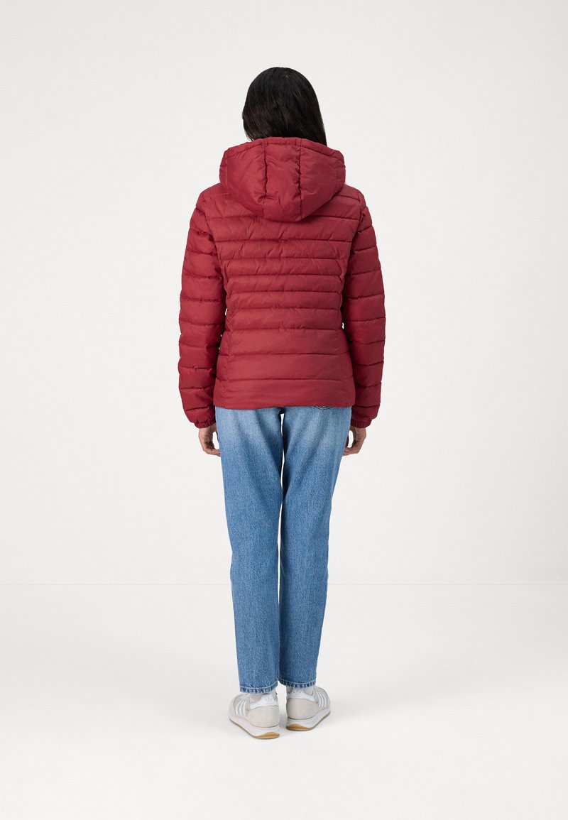 Doudoune rouge avec coutures horizontales, capuche attachée, poignets élastiques et taille cintrée. Associée à un jean en denim bleu et des baskets blanches.