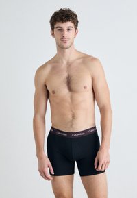 Čierne boxerky s mäkkým pásom, s temne vínovým štítkom s embosovaným nápisom "Calvin Klein" v bielej farbe, priliehavý strih.