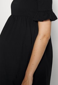Gros plan d'une personne portant une robe noire avec des manches courtes à volants, montrant le bras et le haut du torse.