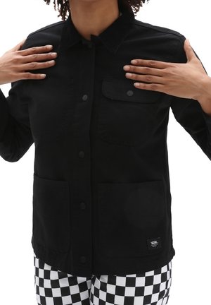 Personne portant une veste noire à boutons avec plusieurs poches et un pantalon à carreaux noir et blanc, les mains touchant le col de la veste.