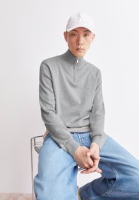 Grauer Zip-Up-Pullover mit hohem Kragen, gerippten Bündchen und kleinem Logo. Kombiniert mit hellblauen Jeans. Weiße Baseballmütze rundet den Look ab.