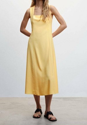 Żółta midi sukienka z szerokimi ramiączkami i kwadratowym dekoltem, o gładkiej fakturze i prostym kroju. Noszona z czarnymi płaskimi sandałami.
