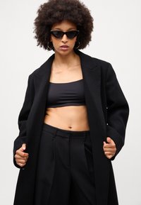 Sort uldfrakke med en skræddersyet silhuet, båret over en sort crop top. Par mørke katteøje solbriller og højtaljede sorte bukser.