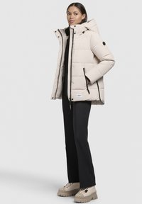 Puffermantel in heller Beige mit einem Reißverschluss vorne, Kapuze und schwarzen Akzenten. Das Modell kombiniert ihn mit schwarzen Hosen und klobigen beigen Stiefeln.
