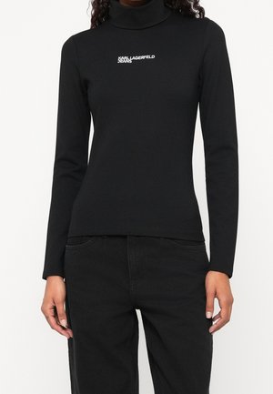 Personne portant un col roulé noir à manches longues avec le texte blanc "KARL LAGERFELD JEANS" centré sur la poitrine et un pantalon noir.