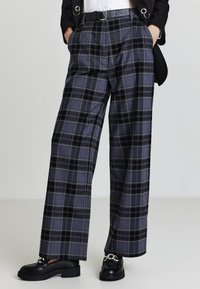 Graue kariert weite Hose mit hoher Taille, versehen mit schwarzen Akzenten und seitlichen Taschen, kombiniert mit schwarzen Slippern.