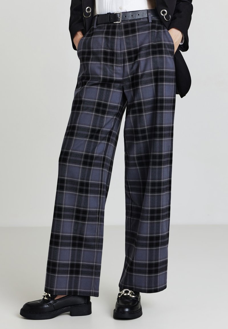 Graue kariert weite Hose mit hoher Taille, versehen mit schwarzen Akzenten und seitlichen Taschen, kombiniert mit schwarzen Slippern.