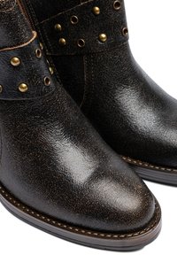 Botines de cuero marrón con acabado texturizado, detalles en latón y tachuelas decorativas de metal a lo largo de la correa, con punta redondeada.