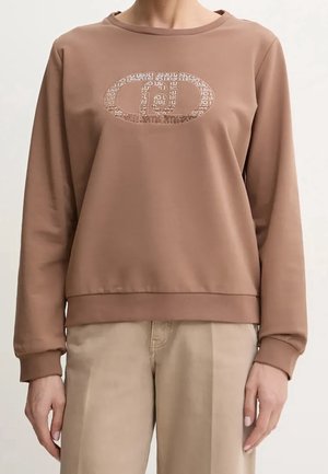 Femme portant un sweat-shirt marron à manches longues avec un logo ovale brillant et à motifs sur la poitrine, et un pantalon beige clair.