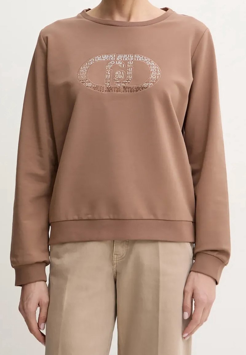 Femme portant un sweat-shirt marron à manches longues avec un logo ovale brillant et à motifs sur la poitrine, et un pantalon beige clair.