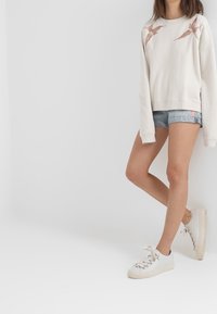Sweatshirt beige clair avec des imprimés d'oiseaux sur les épaules, associé à un short en jean. Le modèle porte des baskets blanches avec des détails en dentelle.