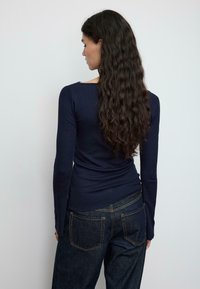 Temno modra majica z dolgimi rokavi in oprijeto zasnovo, rebrasto teksturo ter izrezom v obliki črke U, v kombinaciji s temnimi jeans hlačami z dvema zadnjima žepoma.