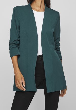 Femme portant un blazer ouvert bleu sarcelle, un haut blanc et un pantalon noir, avec les manches légèrement retroussées et des bijoux minimalistes aux mains.