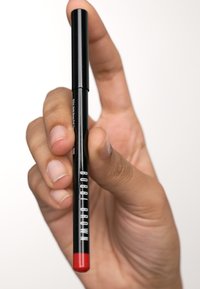 Crayon à lèvres noir tenu entre des doigts, avec une finition brillante, une pointe rouge et une écriture blanche "BOBBI BROWN" sur le côté.