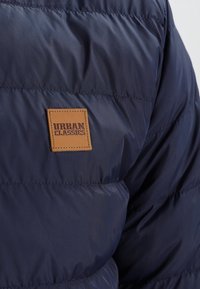 Granatowa pikowana kurtka z kwadratową brązową skórzaną łatą z napisem "Urban Classics" wszytą na plecach.