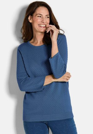 Donna con capelli castani di lunghezza media che sorride, indossando un maglione blu texture e jeans blu, posando su uno sfondo bianco.