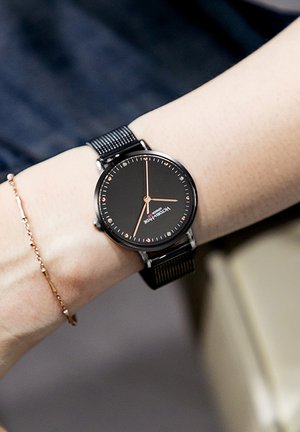 Crni sat s crnim mrežastim remenom, okruglo lice, ružičasto zlatni akcenti, minimalistički dizajn i jasni oznake sati, nošen na zapešću.