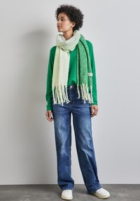 Grøn langærmet top, lys grøn og mørkegrøn mønstret tørklæde med frynser, løse blå jeans og hvide sneakers på en model.