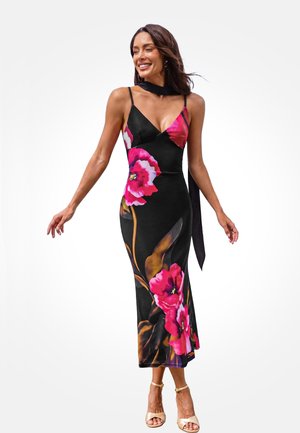 Woman wearing a black sleeveless dress with large pink floral patterns, beige ankle-strap heels, and a black scarf around her neck.