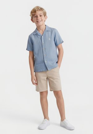 Blond boy standing, wearing light blue short-sleeve button-up shirt, beige shorts, and white sneakers, smiling with hand in pocket.
