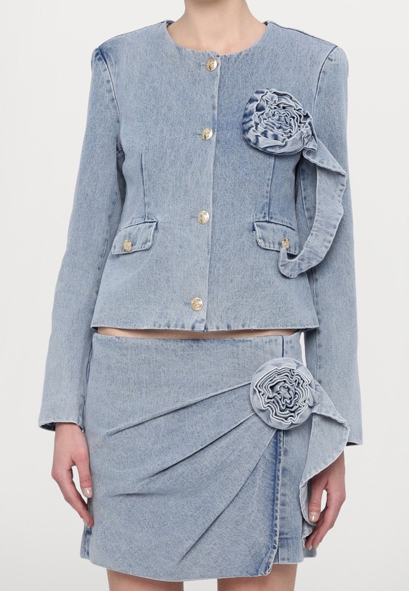 Ensemble veste en denim bleu clair et jupe assortie avec des boutons dorés et des détails en tissu en forme de rosette sur la poitrine et la hanche, porté par une personne.