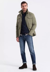 Giacca verde oliva con colletto alto, tasche a quadrifoglio, indossata sopra un maglione blu navy e una camicia a scacchi. Abbinata a jeans blu e stivali marroni.