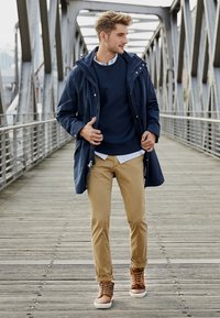 Marine parka met capuchon over een marine sweatshirt, beige chinos en bruine veterschoenen. Outfit gelaagd met een wit shirt. Buitenomgeving op een brug.