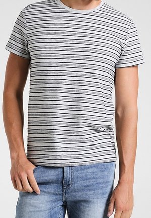 Mężczyzna ubrany w białą koszulkę z krótkim rękawem w cienkie czarne poziome paski oraz jasnoniebieskie dżinsy, stoi z rękami swobodnie opuszczonymi wzdłuż ciała.