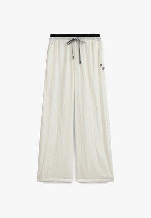 Pantalones de pierna ancha color beige claro con cintura elástica negra y cordón negro, con pequeños parches de logotipo en el lado derecho.