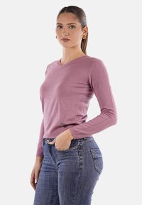 Maglione rosa chiaro a scollo a V realizzato in tessuto lavorato a maglia, con maniche lunghe e un dettaglio con bottone decorativo all'orlo. Indossato con jeans blu.
