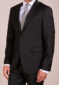 Svart skräddarsydd blazer med smala slag, två knappar och fickor fram. Bärs över en vit skjorta och grå slips. Slät tygliknande textur.