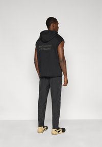 Μαύρο αμάνικο γιλέκο με κουκούλα και λογότυπο στην πλάτη, σε συνδυασμό με σκούρο υφασμάτινο παντελόνι και μαύρα παπούτσια με ταμπά λεπτομέρειες.