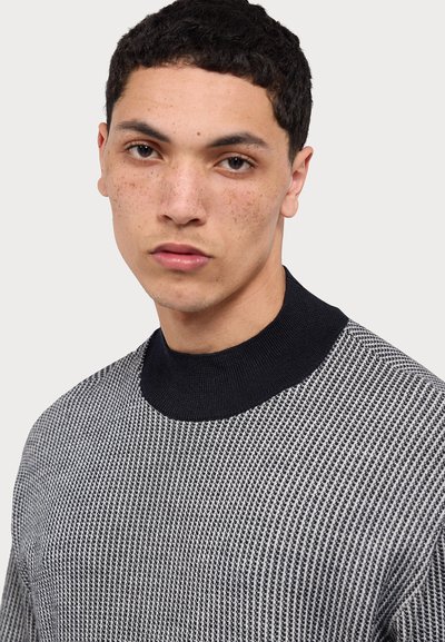 Maglione a maglia con colletto nero, presenta un motivo testurizzato grigio e bianco. Vista ravvicinata dei dettagli del colletto e della parte superiore del torace.