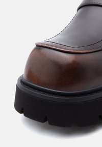 Botte en cuir marron avec un bout arrondi, comprenant un détail cousu sur le col et une semelle épaisse en caoutchouc noir texturée.