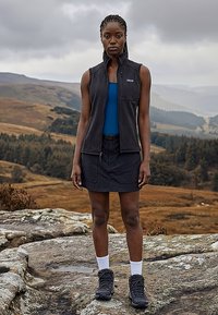 Γυναίκα που φοράει μαύρο γιλέκο, μπλε μπλουζάκι, μαύρη φούστα, ορειβατικά μποτάκια και λευκές κάλτσες, στέκεται σε βραχώδες έδαφος με λόφους στο βάθος κάτω από συννεφιασμένο ουρανό.