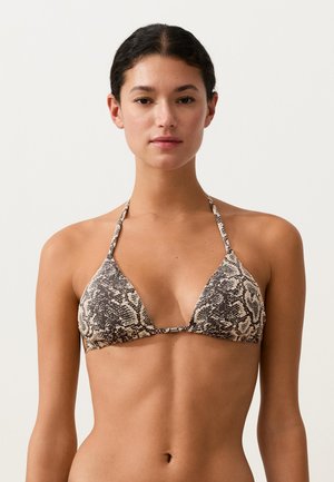 Junge Frau trägt ein braun-beiges Bikinioberteil mit Schlangenmuster und Neckholder-Trägern, steht vor einem schlichten hellen Hintergrund.