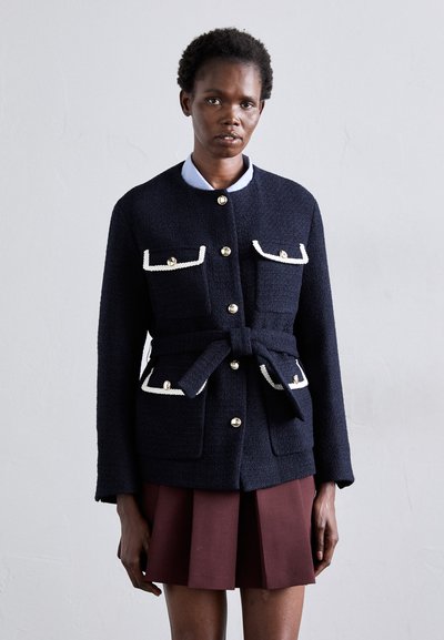 Marineblaue, strukturierte Jacke mit weißen Taschenakzenten und goldenen Knöpfen; hat eine taillierte Form und einen runden Ausschnitt. Getragen über einem hellblauen Hemd.