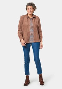 Giacca in suede marrone con bottoni a pressione, abbinata a un top lavorato a maglia multicolore, jeans skinny blu e stivali marroni con lacci.